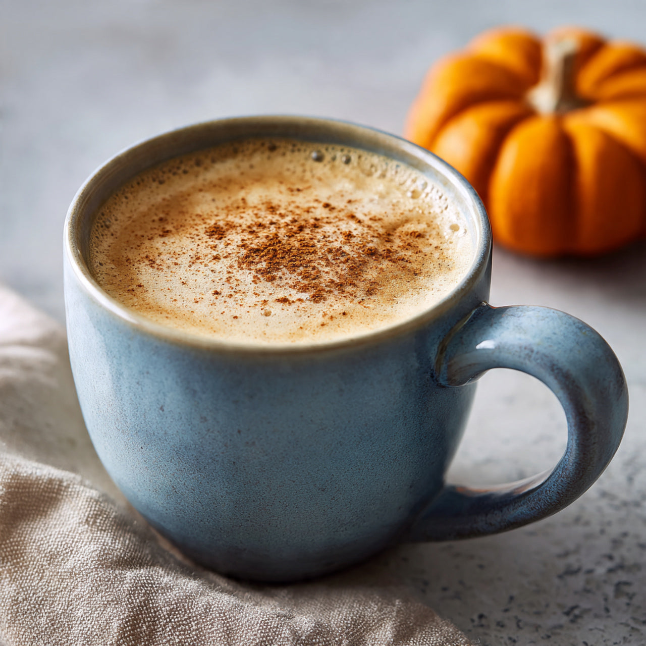 Blue mug of coffee with pumpkin spice on a stone surface with a small pumpkin in the background.