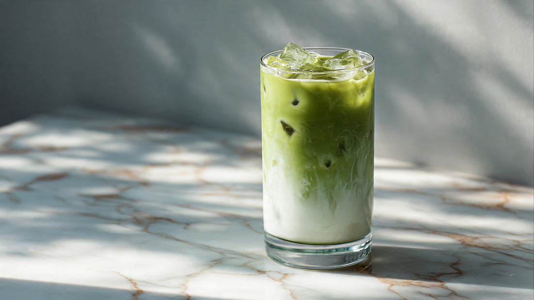 Iced matcha latte in clear glass on pale stone table with bamboo whisk accent in soft natural light