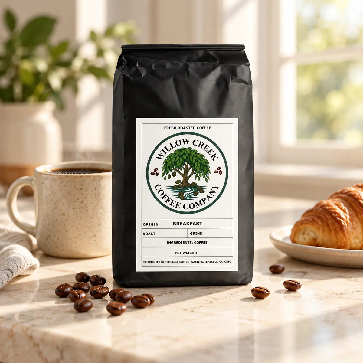 Willow Creek Coffee Company Breakfast Blend bag beside a ceramic mug of black coffee and a golden croissant on a sunlit kitchen countertop.
