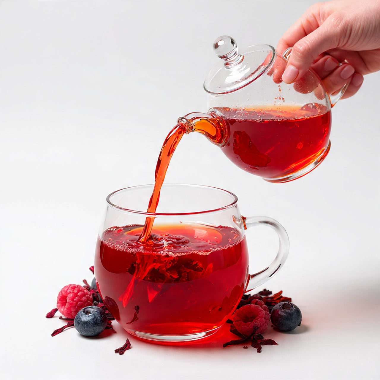 hibiscus berry tea being poured from a glass teapot into a glass mug on a white background.