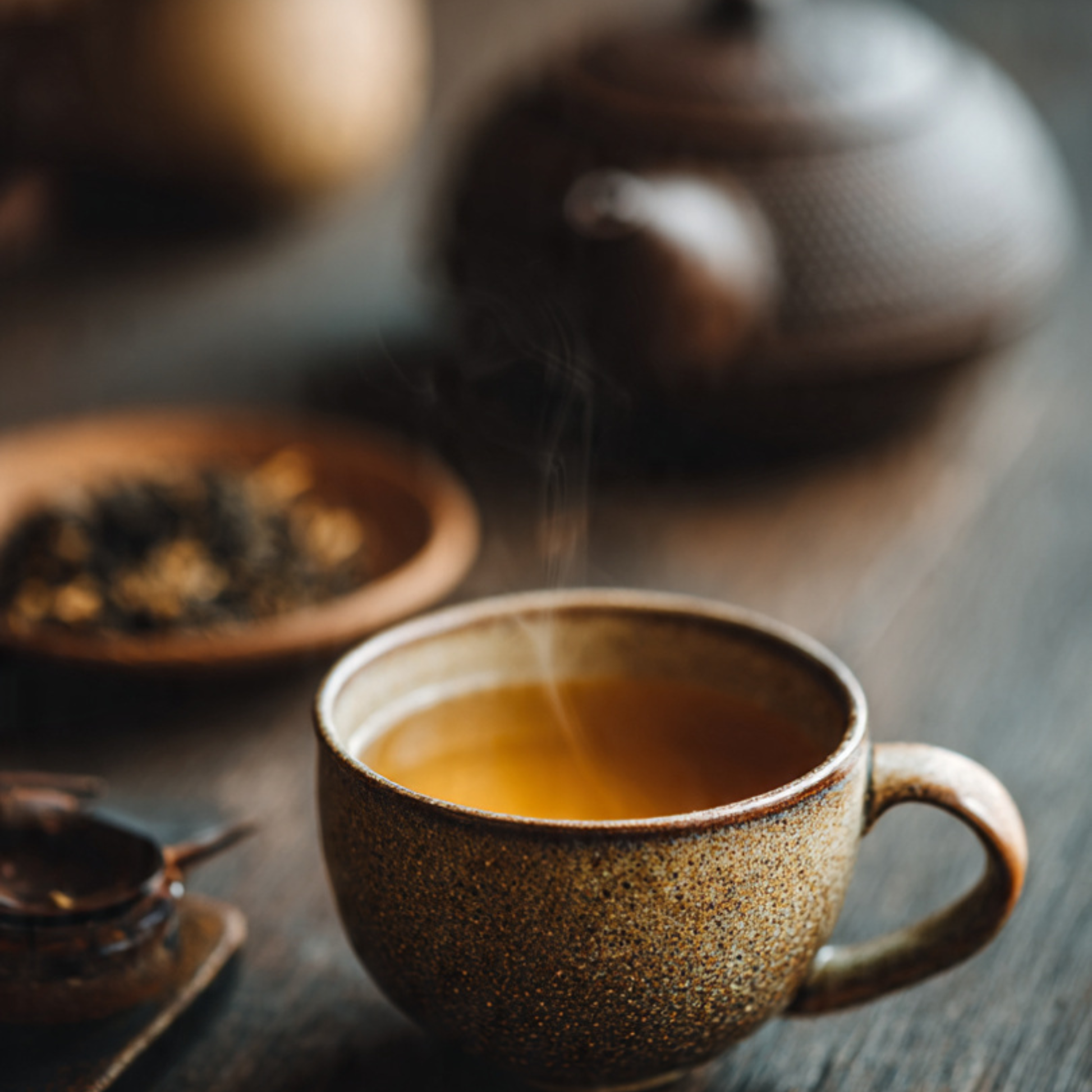 Steaming cup of tea on a wooden table with a teapot and tea leaves in the background.