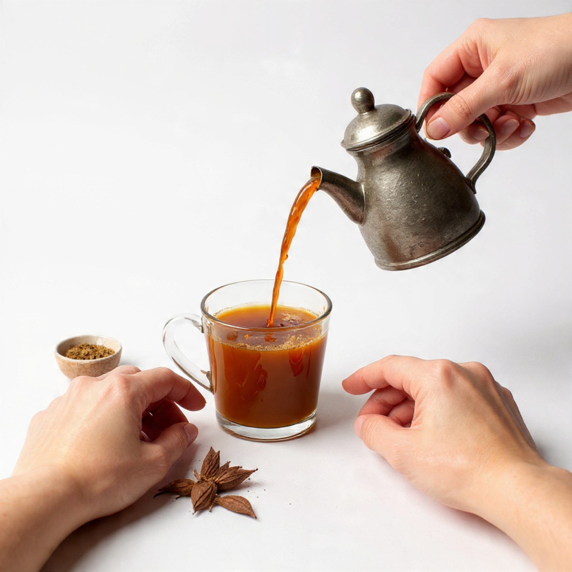 masala-chai tea being poured from a silver teapot into a glass mug on a white surface.
